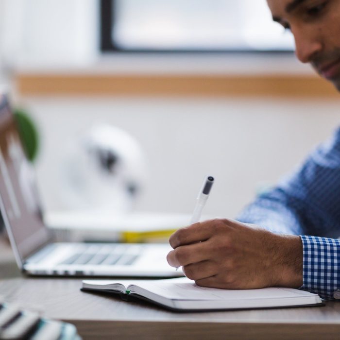 man writing on notebook next to laptop